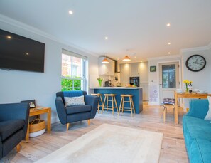 A modern living room with a sofa, a blue armchair, and a coffee table. There is a kitchen area with a bar and stools. - 2 The Lookout - Two Bedroom Apartment, Sleeps 4 (Swanage)