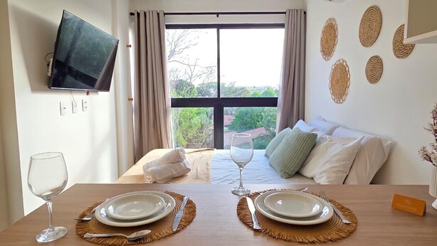 Habitación de hotel con una mesa de comedor para dos personas, una cama con almohadas, un televisor montado en la pared y un amplio ventanal con cortinas.