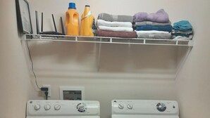 A laundry area with two white front-loading washers and dryers, a shelf above with cleaning supplies and folded towels.