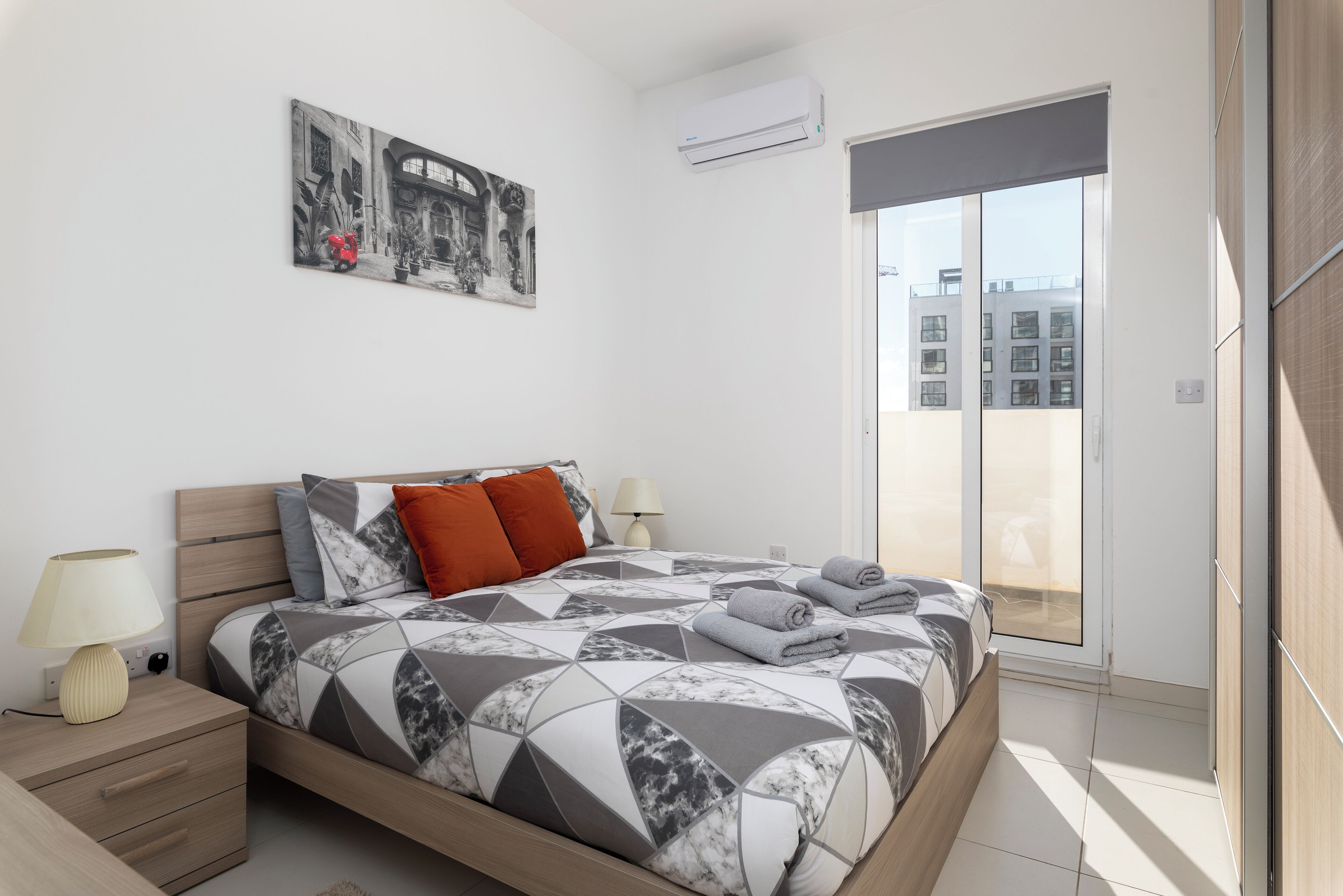 A modern bedroom with a geometric-patterned bedspread, a nightstand with a lamp, a wall-mounted air conditioner, and a large window offering a view of buildings.