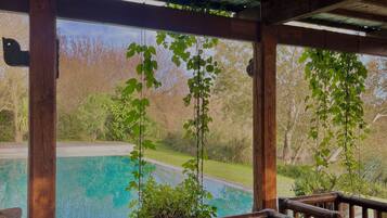 Une terrasse en bois avec une piscine, agrémentée d’une pergola ornée de plantes suspendues et offrant une vue sur un paysage naturel.