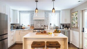 Une cuisine moderne avec des meubles blancs, un îlot central doté de tabourets, un éclairage suspendu et un dosseret orné d’un motif en chevrons.