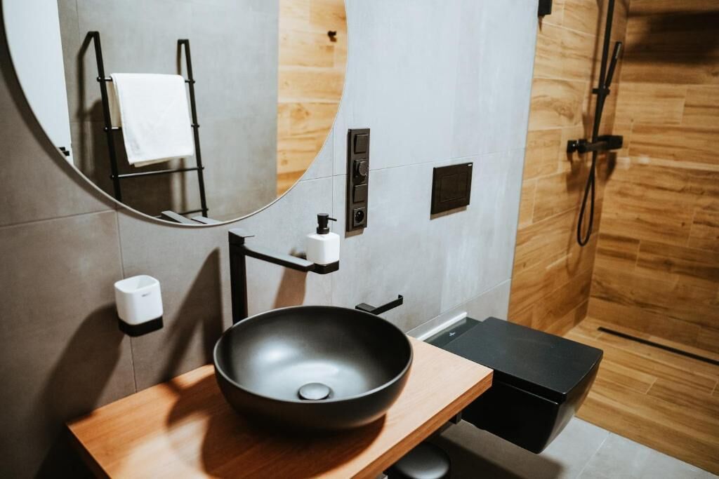 A modern bathroom with a wooden vanity, a black basin, a mirror, a towel rack, and a shower area.