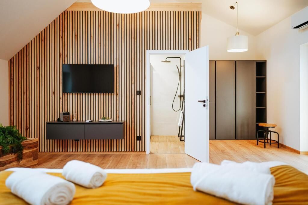 A modern bedroom with a sloped ceiling, a mounted TV, a wooden wall panel, a bathroom with a shower, and a bedside table with towels.