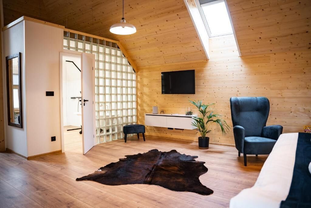 Un interior moderno con techo inclinado, paredes de madera, un gran tapete de cuero, un sofá, una silla, un escritorio con una planta y un televisor montado en la pared.
