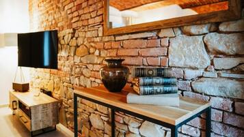 A room with a stone wall, a wooden table with books and a vase, a TV on a stand, and a mirror.