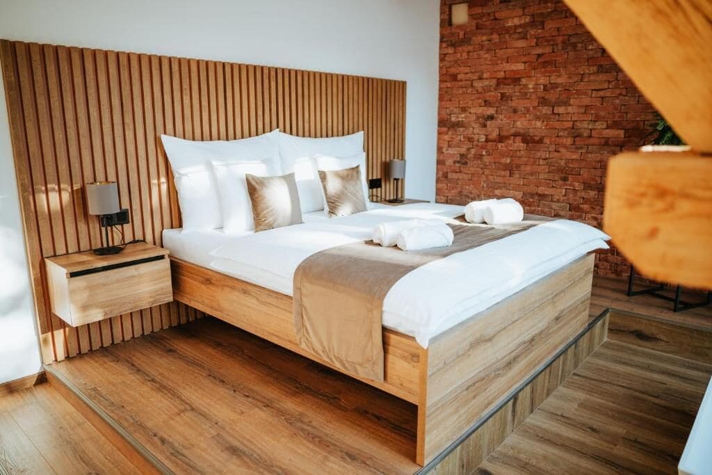 Un dormitorio moderno con cabecera de madera, una cama grande con ropa de cama blanca, una mesita de noche de madera y una pared de ladrillo.