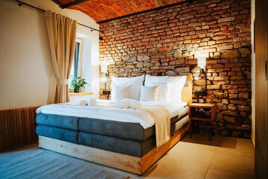Un dormitorio con pared de piedra, una cama con sábanas blancas, cabecera de madera y una mesita de noche con lámpara.