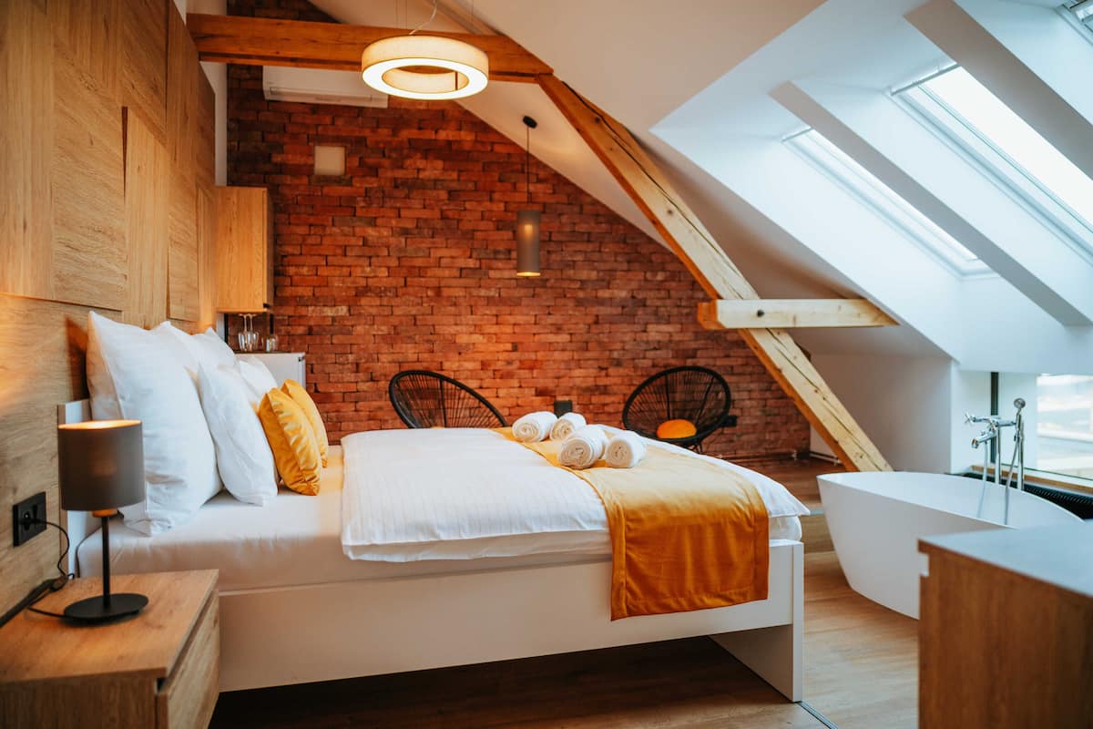 A modern bedroom with a brick wall, a sloped ceiling, a skylight, a bed with white bedding, a bedside table with a lamp, and a bathtub.