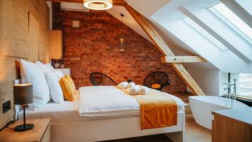 A modern bedroom with a brick wall, a sloped ceiling, a skylight, a bed with white bedding, a bedside table with a lamp, and a bathtub.
