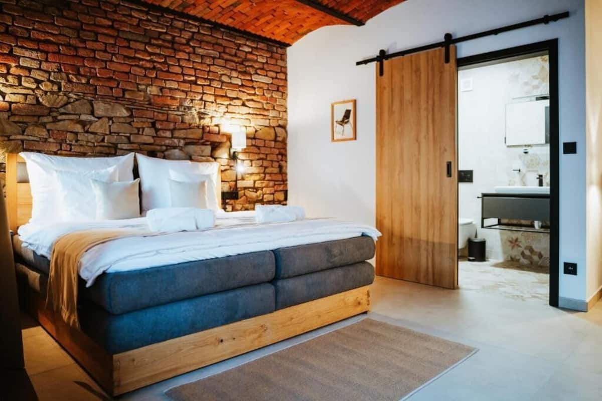 A bedroom with a brick wall, a wooden bed frame, a sliding door, and a bathroom visible through the door.