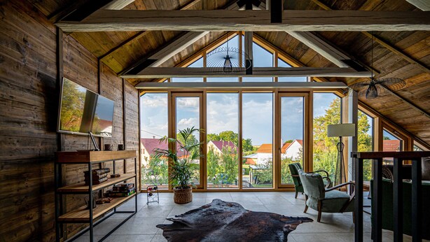 A spacious room with a large window, wooden beams, a leather rug, a TV on a shelf, and a chair. - Galerie mlýn Žeraviny (Žeraviny)