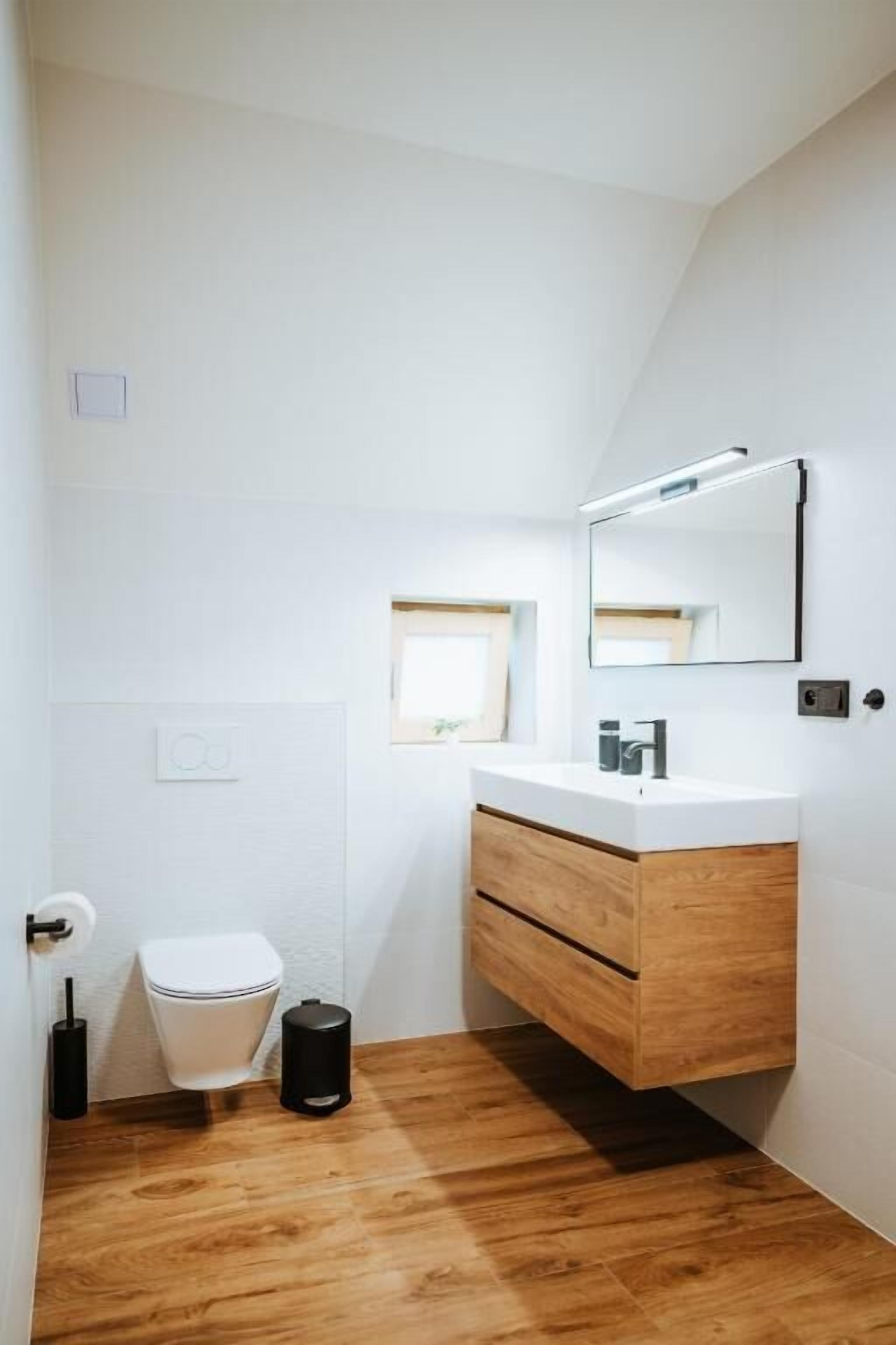 A small, modern bathroom with a wooden vanity, a white toilet, and a small window providing natural light.
