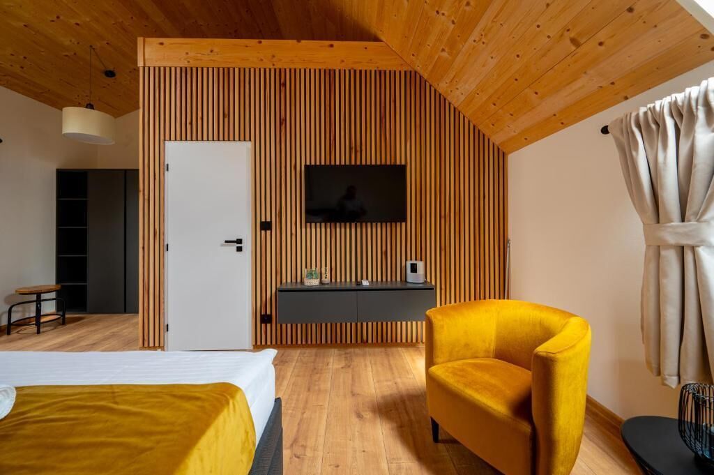 Ein modernes Schlafzimmer mit Holzboden, einem Bett mit gelber Bettdecke, einem Schreibtisch mit Computer und einem an der Holzwand montierten Fernseher.