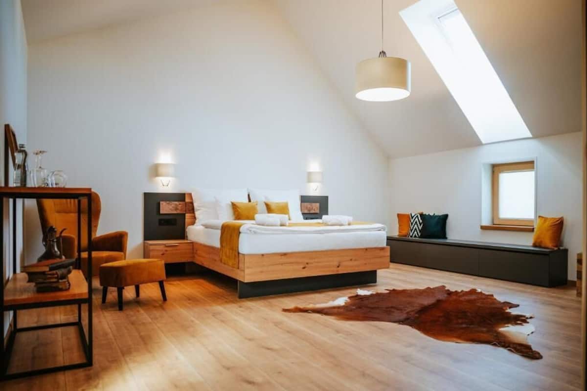 A modern bedroom with a sloped ceiling, a large bed, a wooden side table, a chair, and a leather rug.