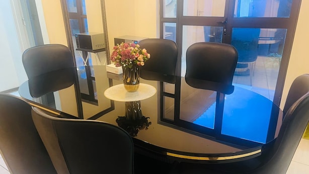 Un'area pranzo moderna con un tavolo in vetro circondato da sedie nere, un piccolo tavolino tondo con un vaso di fiori e vista attraverso la porta in vetro.