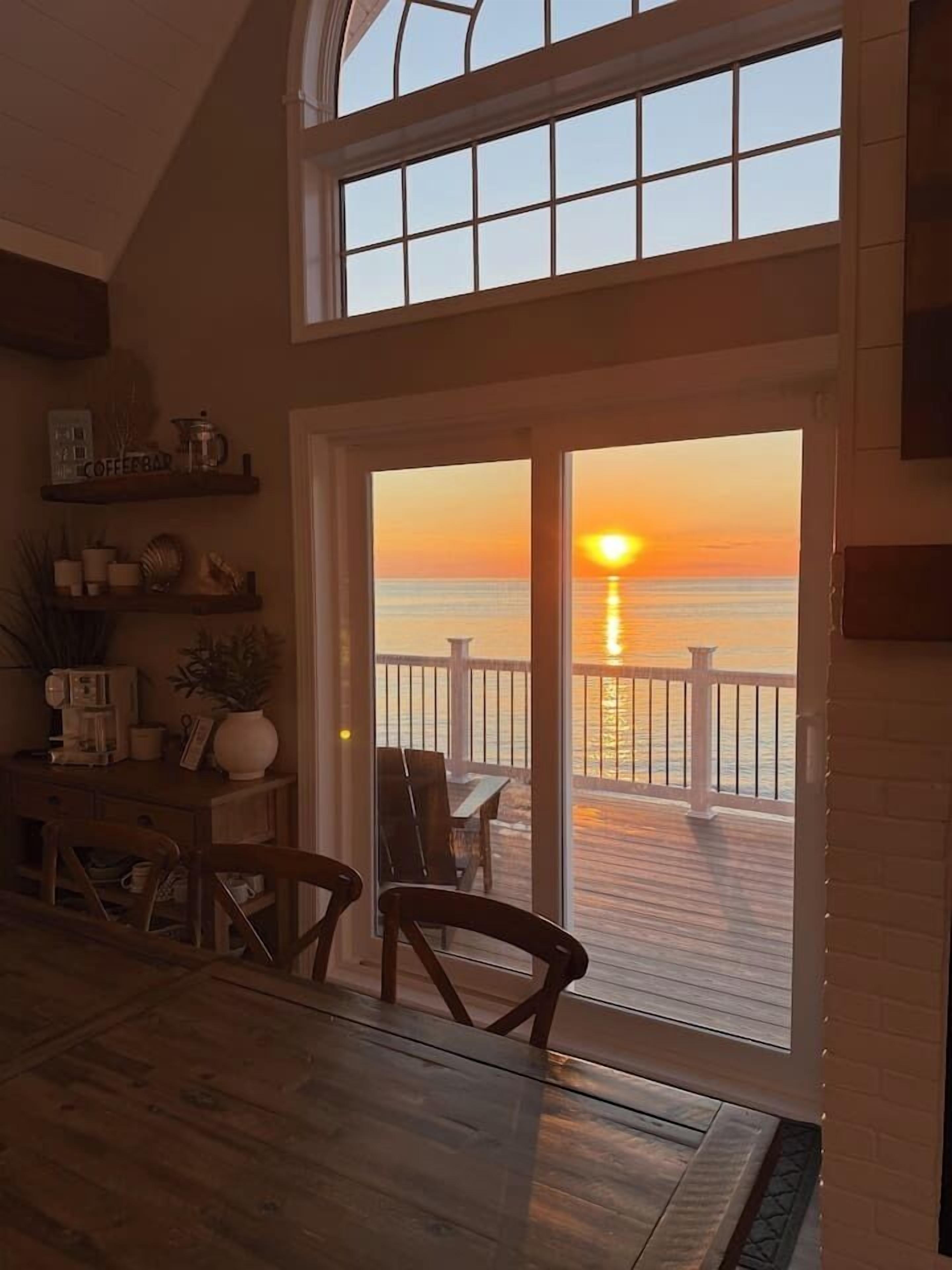 Un espace repas offrant une vue sur le coucher de soleil sur l’océan, une terrasse en bois et une porte vitrée donnant sur l’extérieur.