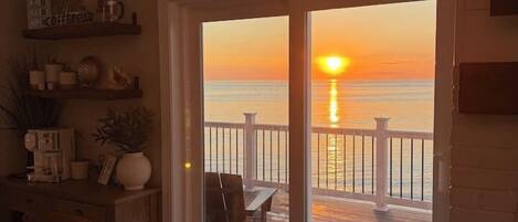 Un espace repas offrant une vue sur le coucher de soleil sur l’océan, une terrasse en bois et une porte vitrée donnant sur l’extérieur.