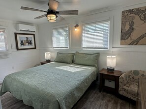 A bedroom with a bed, two bedside tables, a ceiling fan, and a window with blinds. - Doggy Paddle: Pet-Friendly Couple's Retreat (Ocracoke)