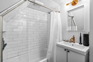 A modern bathroom with white subway tiles, a glass-enclosed shower, a white sink with a gold faucet, and a black electric toothbrush charger. - Allentown Cottage: Walk to Arts District, Eclectic Food, and Buffalo Nightlife (Buffalo)
