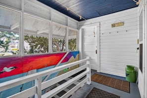 A covered porch with a colorful mural, a white door, and a green trash can.
