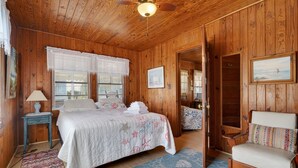 Habitación con paneles de madera, una cama, una silla, una lámpara y un ventilador de techo.