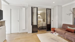 Ein modernes Wohnzimmer mit Sofa, Couchtisch und Fernseher. Eine Tür führt zu einem Schlafzimmer mit Bett und Nachttisch.