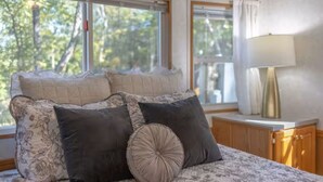 Une chambre à coucher avec un lit, des oreillers, une table de chevet, une lampe et une fenêtre donnant sur des arbres.