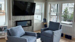 A living room with two grey armchairs, a flat-screen TV mounted on a stone wall, and large windows offering a view of the outside. - Brand New! Enjoyable 5-bedroom house with AC in Wells Beach (Wells)