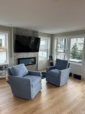 Ein Wohnzimmer mit zwei grauen Sesseln, einem an einer Steinwand montierten Flachbildfernseher und großen Fenstern mit Blick hinaus.