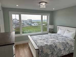 A bedroom with a large window, a bed with a patterned bedspread, a bedside lamp, and a view of houses and greenery outside. - Brand New! Enjoyable 5-bedroom house with AC in Wells Beach (Wells)
