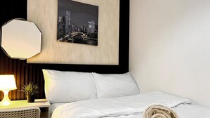 Ein ordentlich bezogenes Bett mit weißen Leinentüchern, ein aufgerolltes Handtuch, ein gerahmtes Stadtbild, eine moderne Lampe und ein kleiner Topfpflanze auf einem Nachttisch.