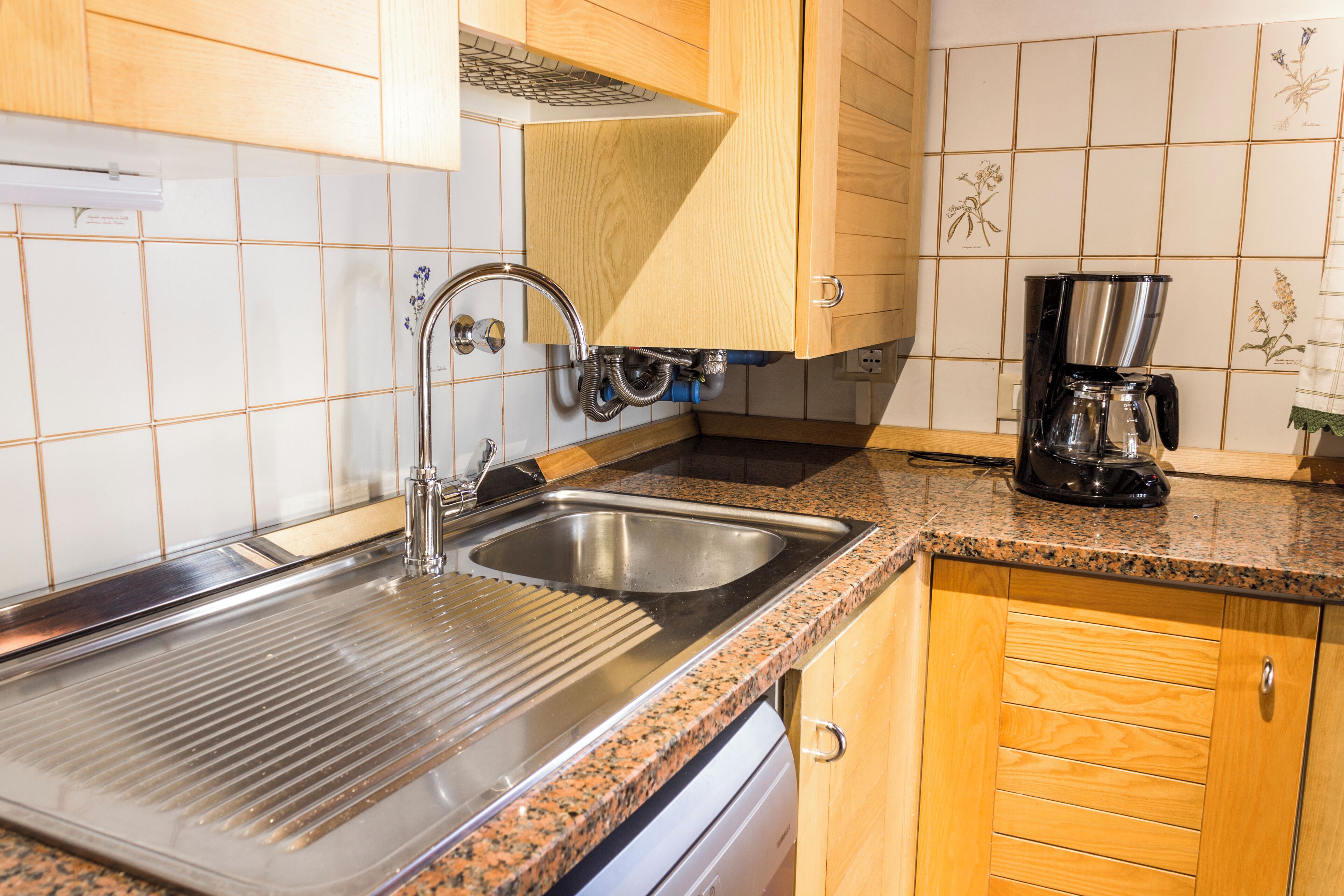 Una cocina con un lavamanos de acero inoxidable, armarios de madera, una cafetera y un salpicadero de azulejos.