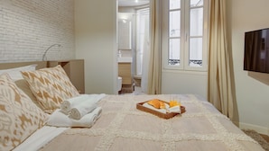 Une chambre d’hôtel avec un mur en briques, un lit agrémenté d’oreillers à motifs, un plateau avec des croissants et du jus d’orange, et un téléviseur fixé au mur.