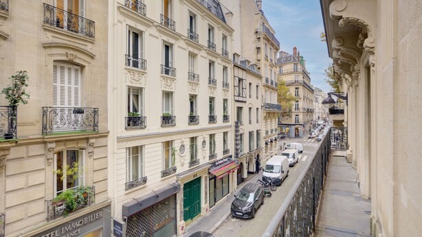 Une rue étroite bordée d’immeubles à plusieurs étages, avec des balcons, des devantures de magasins et des voitures garées.