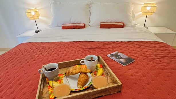 En snyggt bäddad säng med vita lakan och röda kuddar, ett brickbord med croissanter, kaffe och juice, och två sänglampor.
