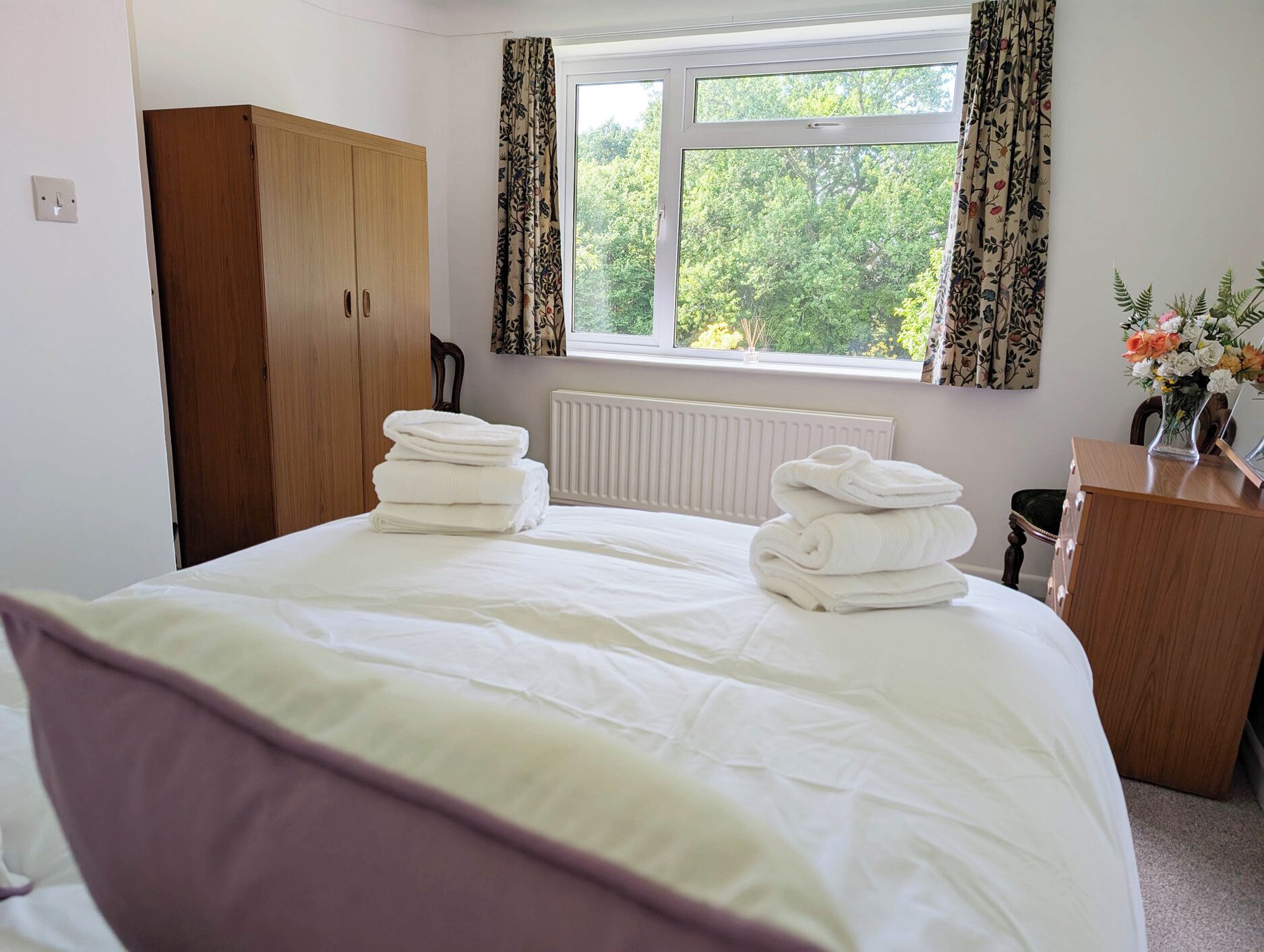 A bedroom with a bed, a wardrobe, a radiator, a window with curtains, and a dresser with a vase of flowers.