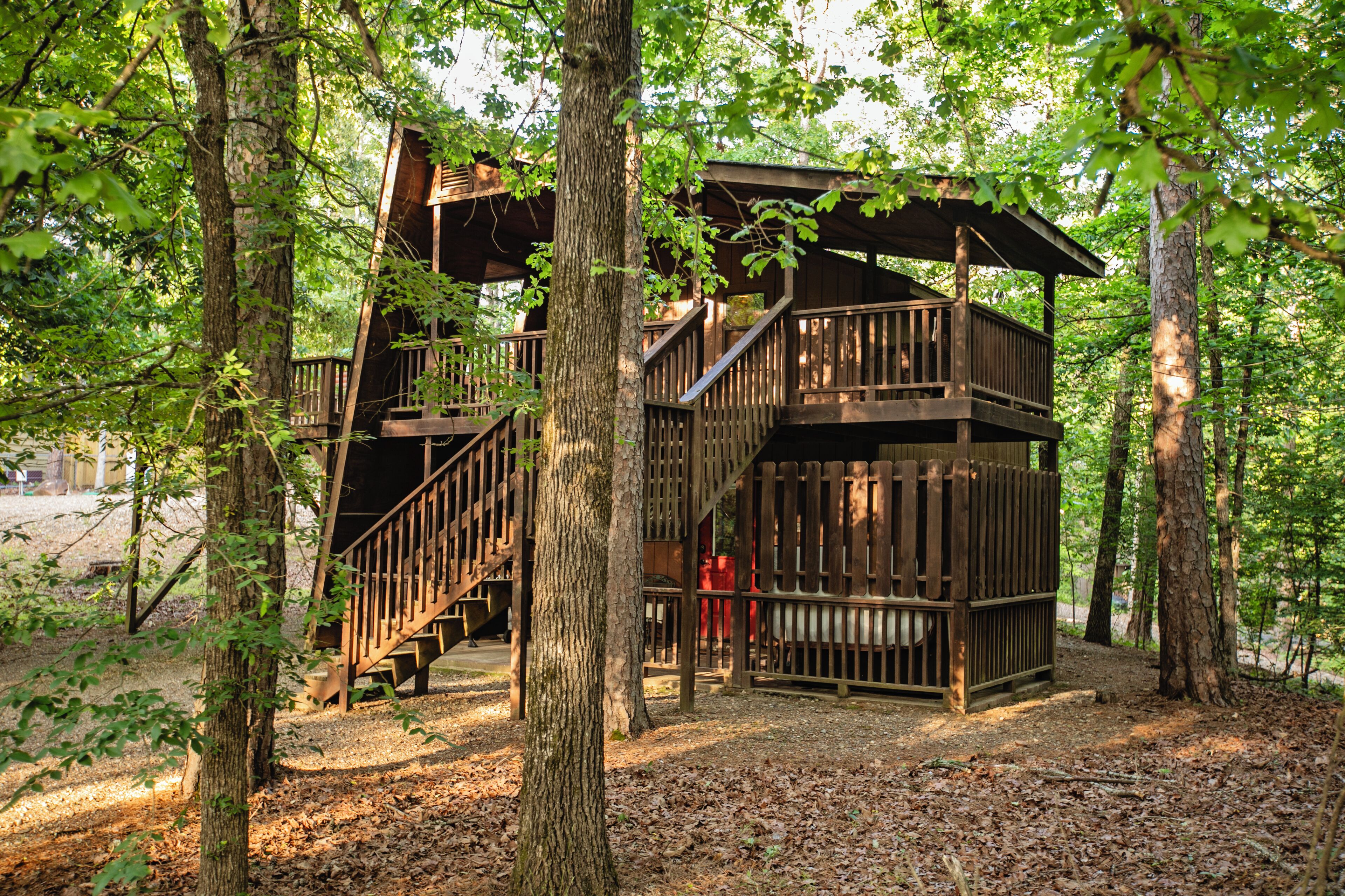 A wooden treehouse with multiple levels and a staircase, surrounded by trees in a forested area.