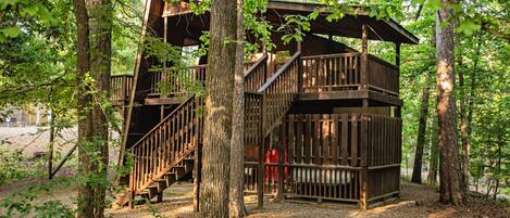 A wooden treehouse with multiple levels and a staircase, surrounded by trees in a forested area.
