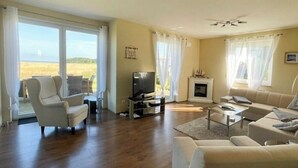 A spacious living room with a sofa, armchair, and a television. There is a fireplace and a large window with curtains. - OstseeWellenrauschen 13 Arrive and feel right at home, (Dranske)