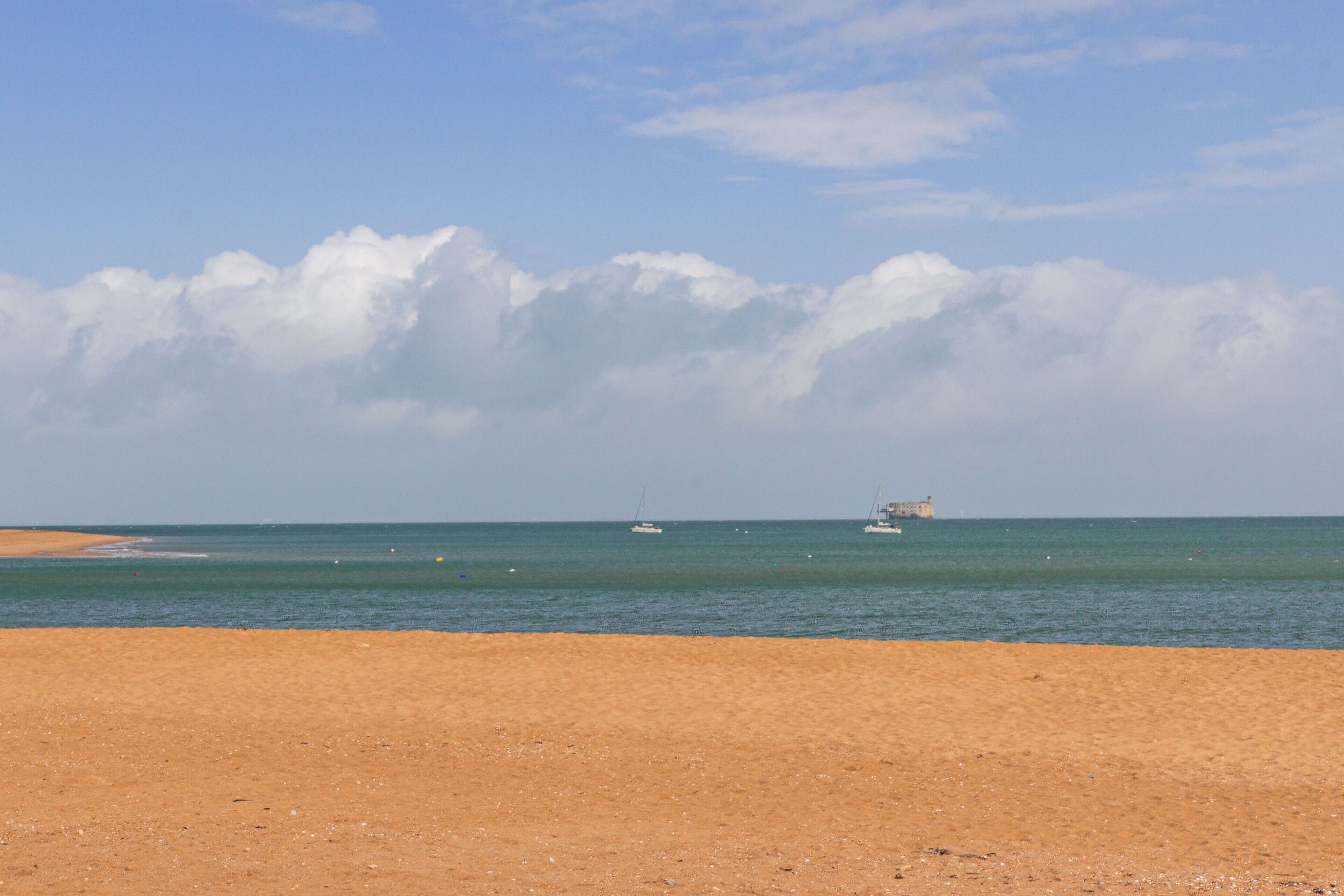 Una playa con arena, un mar tranquilo y algunos barcos a lo lejos.