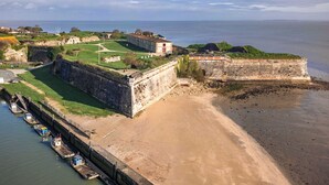 A coastal fortification with a grassy area, a paved walkway, and a row of boats docked along the shore. - The Little House with Its Palm Tree (Dolus-d'Oléron)