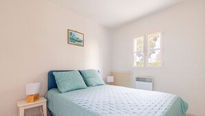 A bedroom with a bed, a nightstand, a wall-mounted lamp, a radiator, and a window. - The Little House with Its Palm Tree (Dolus-d'Oléron)