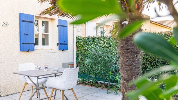 A patio with a table and chairs, a window with blue shutters, and a hedge. - The Little House with Its Palm Tree (Dolus-d'Oléron)