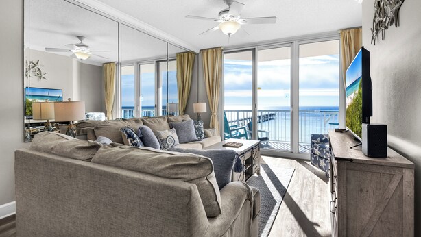 Ein geräumiges Wohnzimmer mit einer großen Couch, einem Couchtisch und einem Fernseher auf einem Holzgestell. Große Fenster bieten Blick auf das Meer.