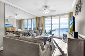 Un salon spacieux avec un grand canapé, une table basse et un téléviseur posé sur un meuble en bois. De grandes fenêtres offrent une vue sur la mer.