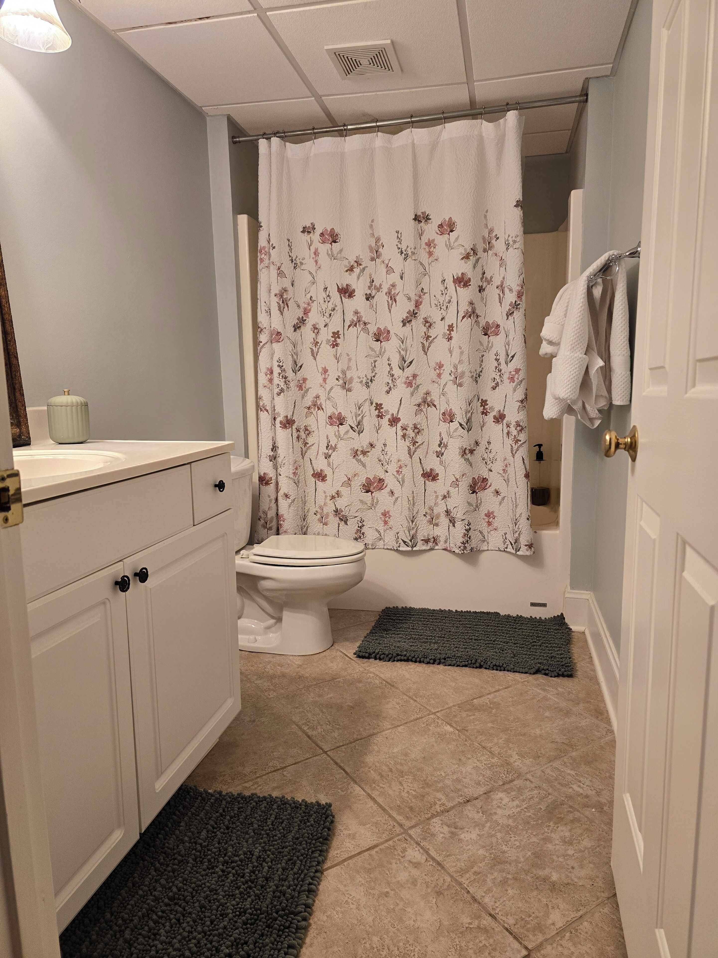 A bathroom with a toilet, a sink, a shower curtain with floral patterns, and two dark mats on the floor.