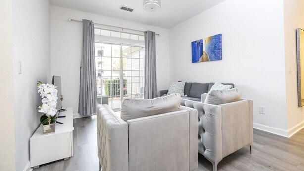 Een moderne woonkamer met een grijze zithoek, een witte bijzettafel met een orchidee, en een groot raam met grijze gordijnen.