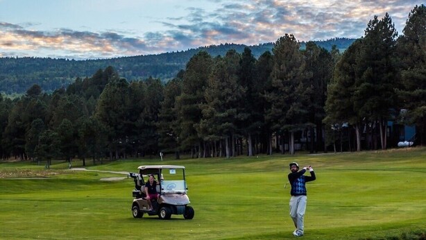 A golf course with a water body in the foreground, a golf cart, and a golfer in mid-swing. - Cozy 2BR Mountain Condo in Angel Fire Near Slopes & Trails. Perfect for families (Angel Fire)