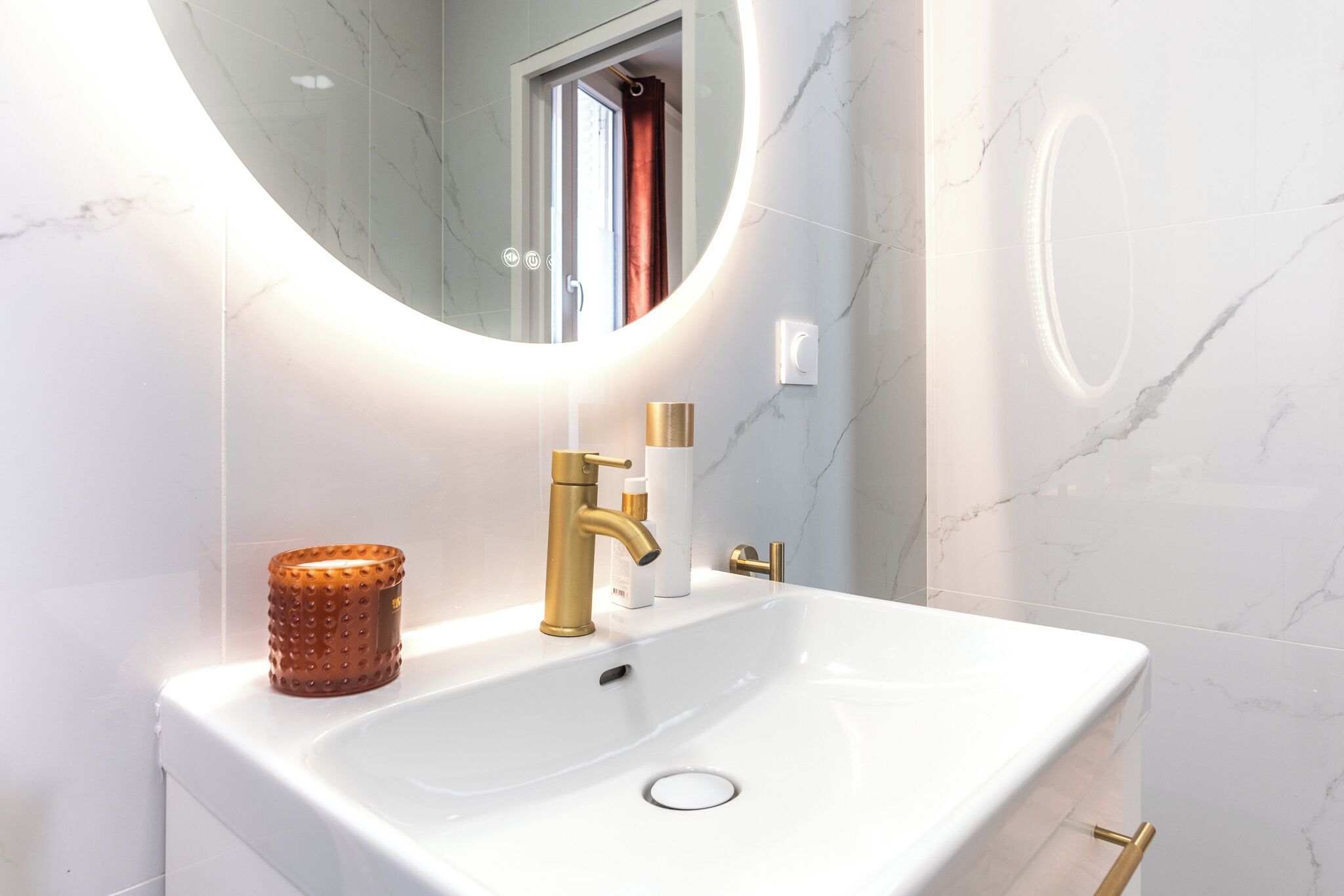 A bathroom with a white sink, gold faucet, and a round mirror with a light.